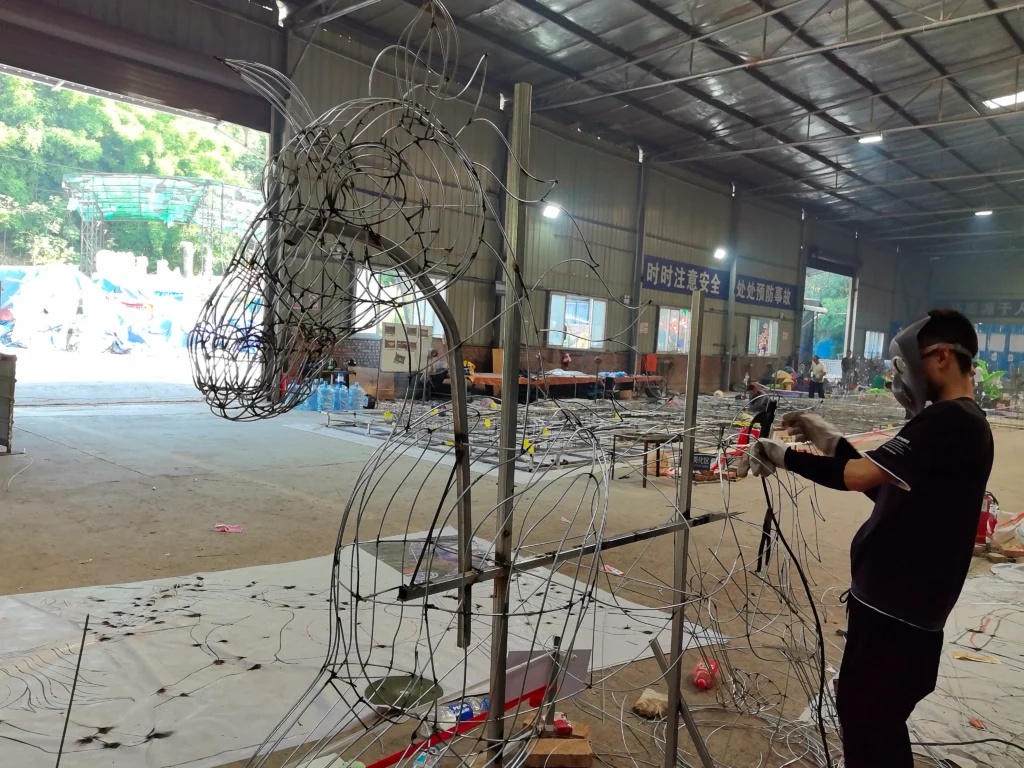 A worker shapes an exposed steel-and-wire horse framework in a workshop.
